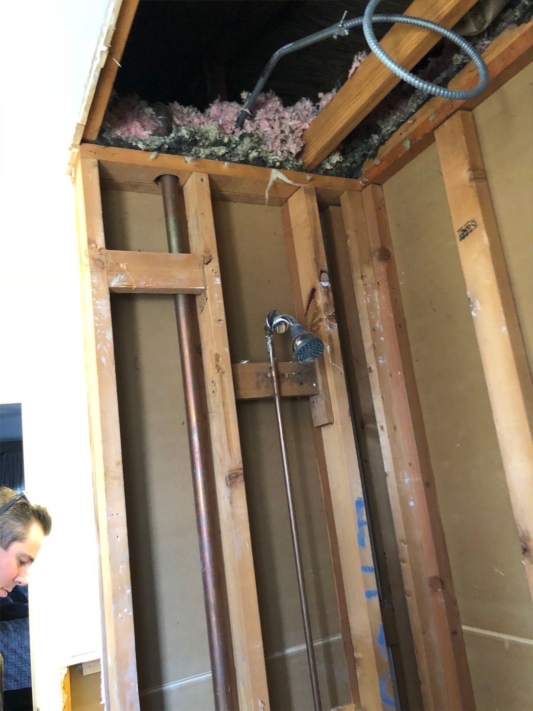 Interior wall framing with exposed plumbing. Insulation visible above. A person stands nearby.