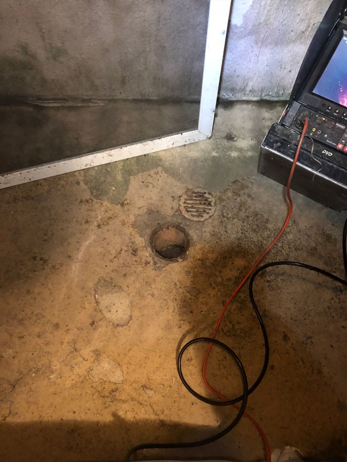 Basement floor with drain and laptop, potentially for inspection or assessment. Concrete floor, orange and black wires.