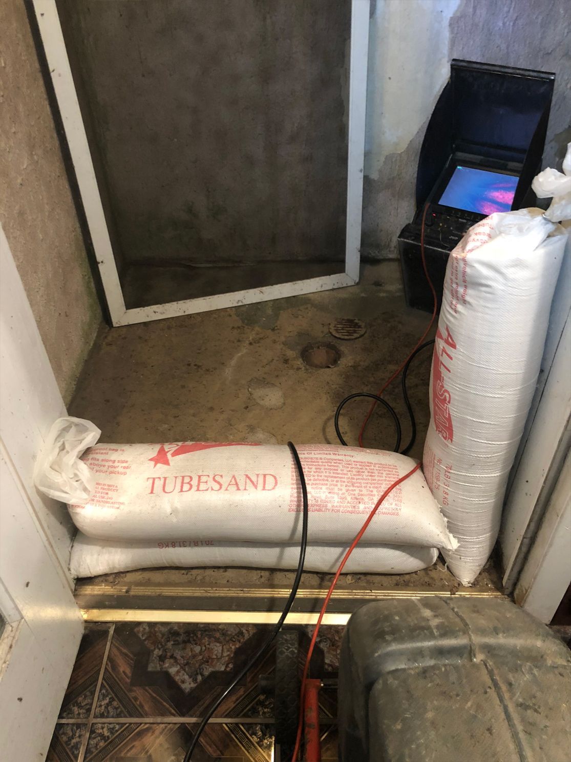 Bags of sand inside a doorway. A welding machine sits behind them, with a screen showing a blue light. Concrete floor.