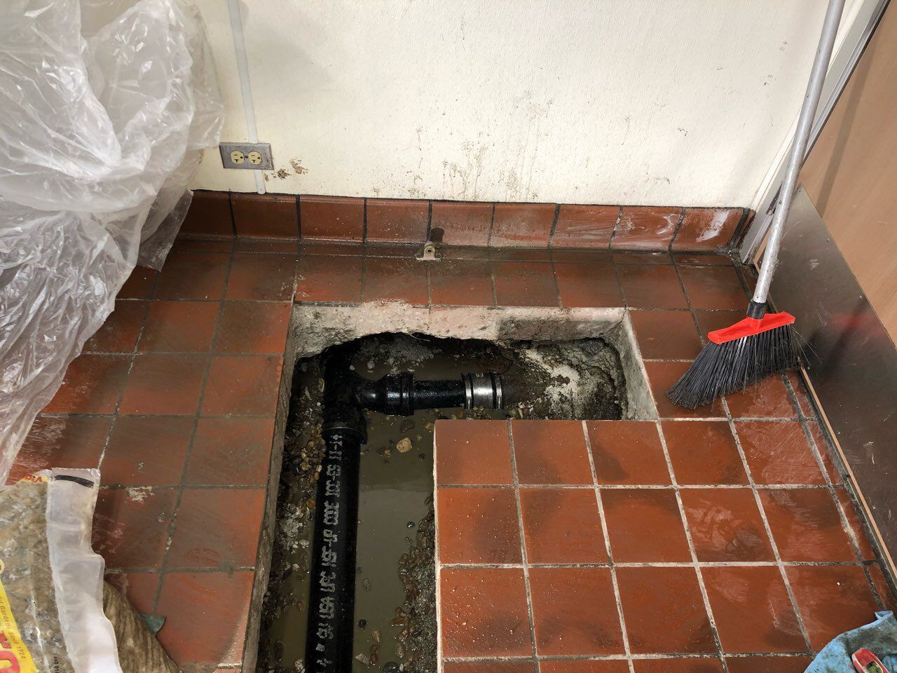 Hole in tiled floor reveals plumbing. Black pipe runs vertically. Reddish tiles surround the opening, next to a wall.