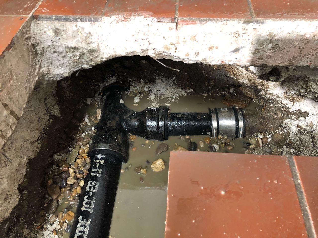 Black pipe fittings in a hole in the floor with water, surrounded by tile and concrete debris.