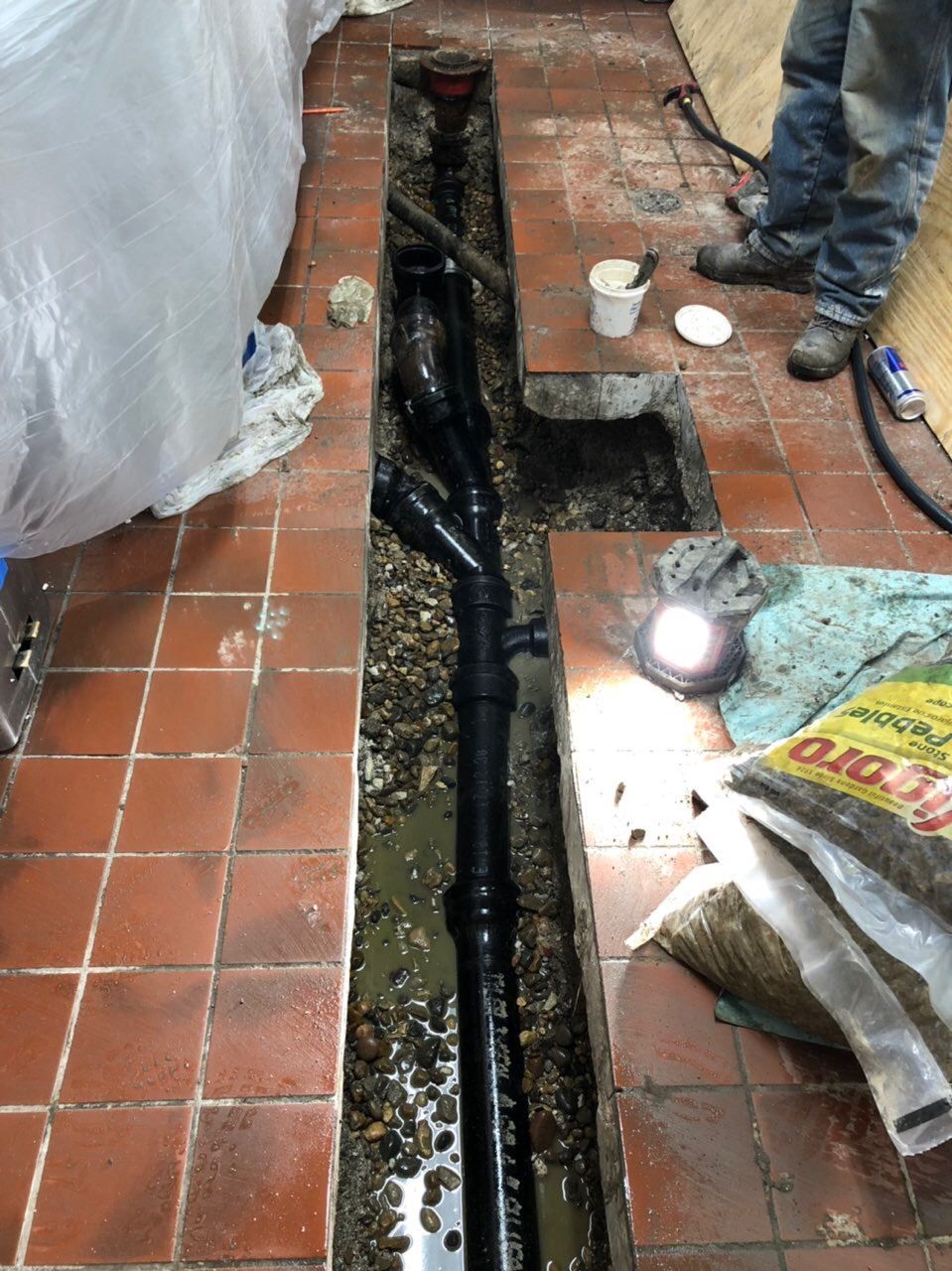 Trench in tiled floor with black pipes; a person's legs are visible nearby.
