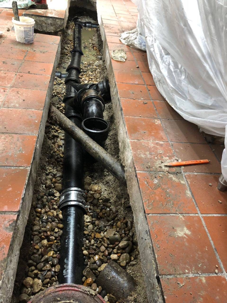 Black plumbing pipes and fittings installed in a narrow trench between red tiles, gravel and a white sheet.