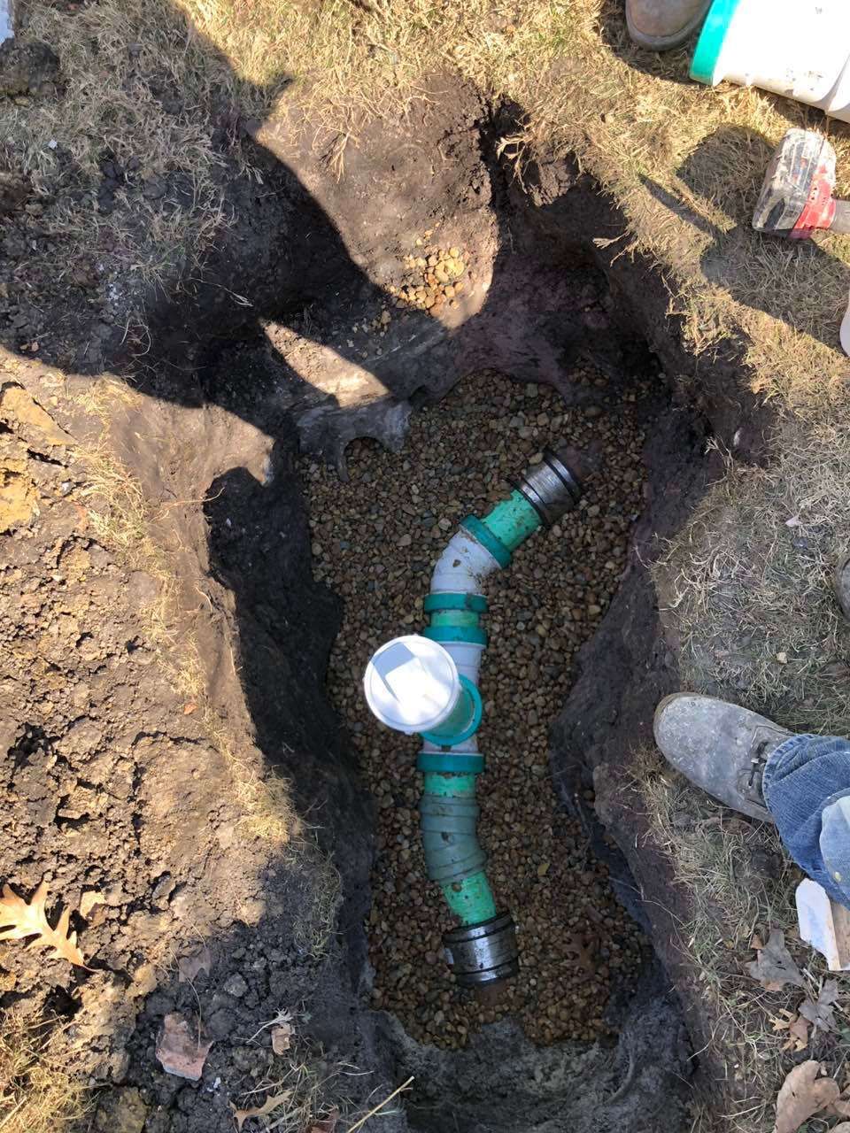Drainage pipe in a gravel-filled trench. Green and gray pipe connections. Someone's feet and dirt are visible.