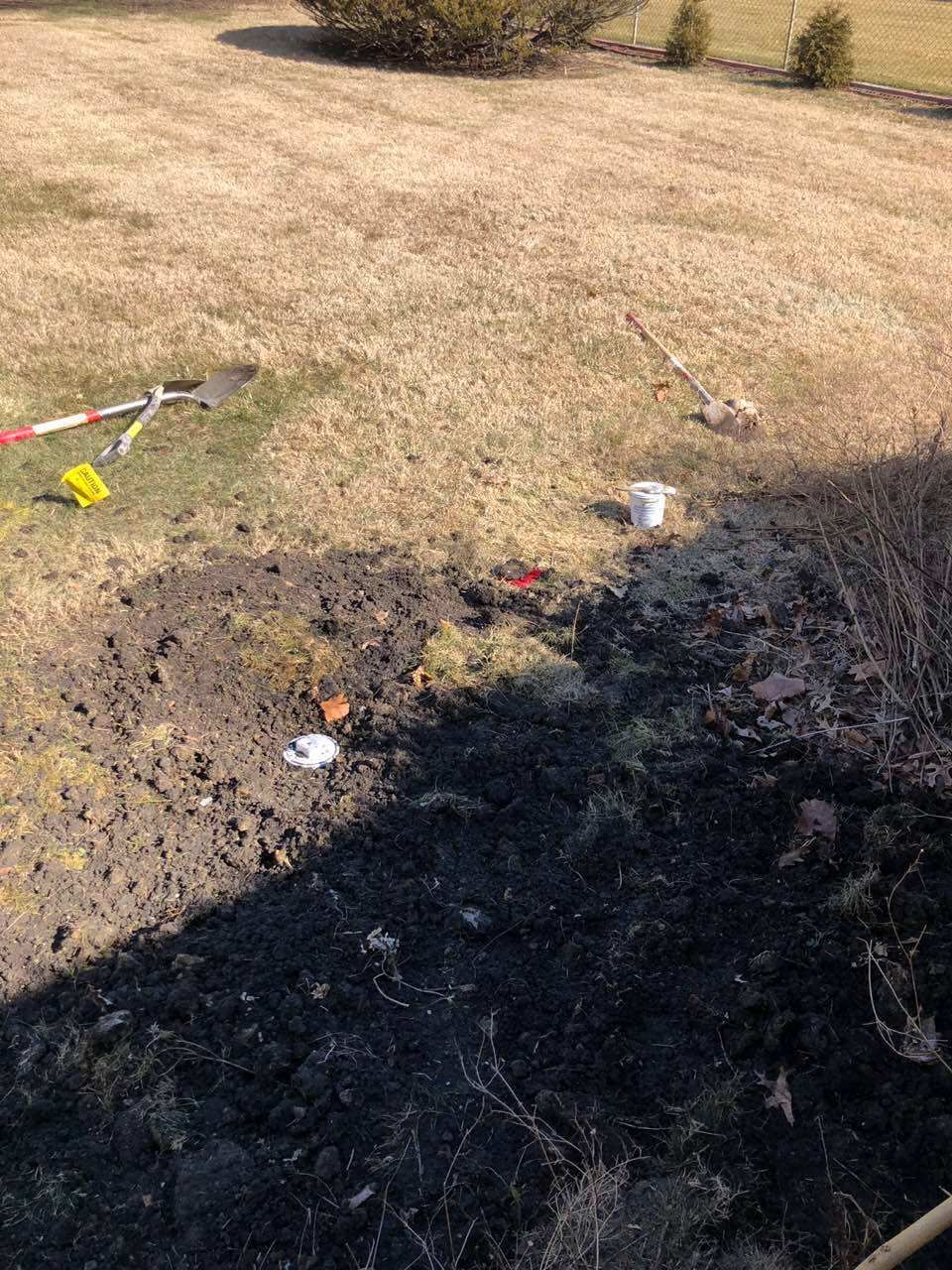 Brown, charred earth with debris, a bucket, and a shovel, in a field of dry grass and small bushes.