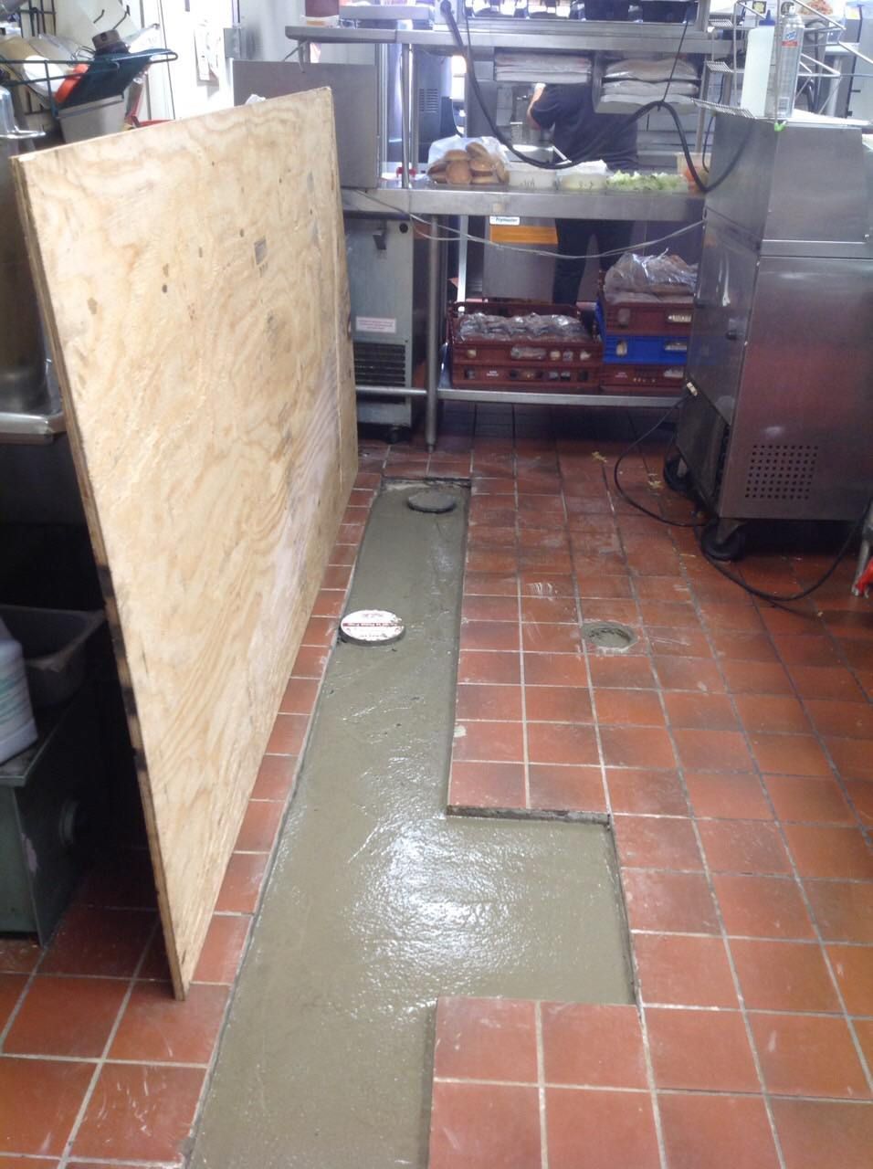 A kitchen floor under repair; a section of tiles removed, concrete poured; plywood leans against a wall.