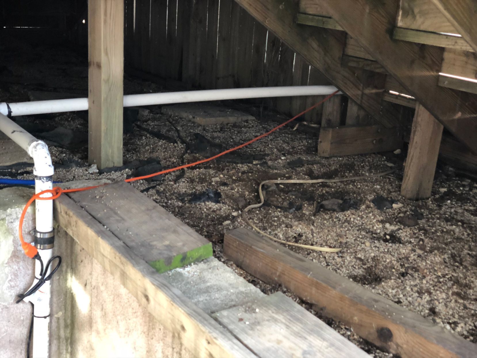 Under-deck view of plumbing, electrical wire, and wood supports in a dark, confined space with debris on the ground.