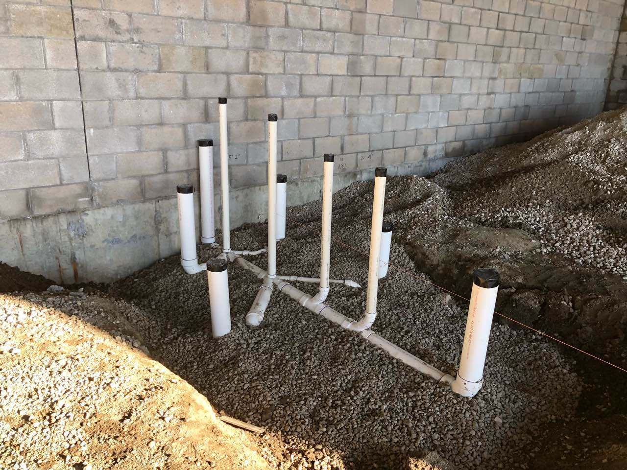 White plumbing pipes, capped, extending from gravel bed near brick wall.