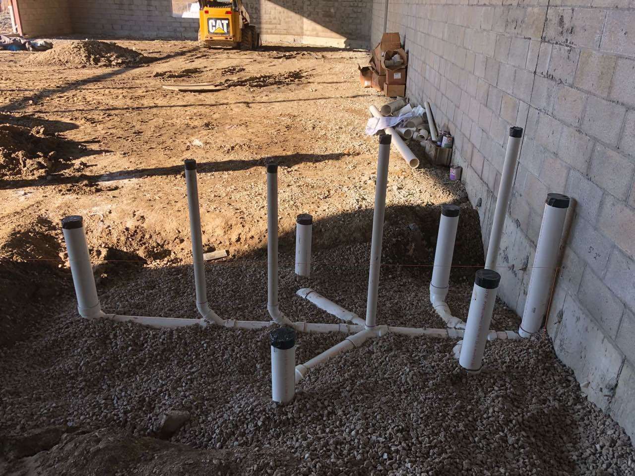 White plumbing pipes on gravel ground next to a brick wall at a construction site.