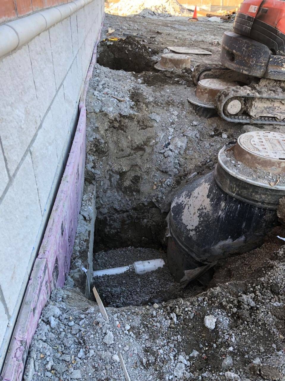Construction site with a deep hole next to a brick building; black pipe and dirt visible.