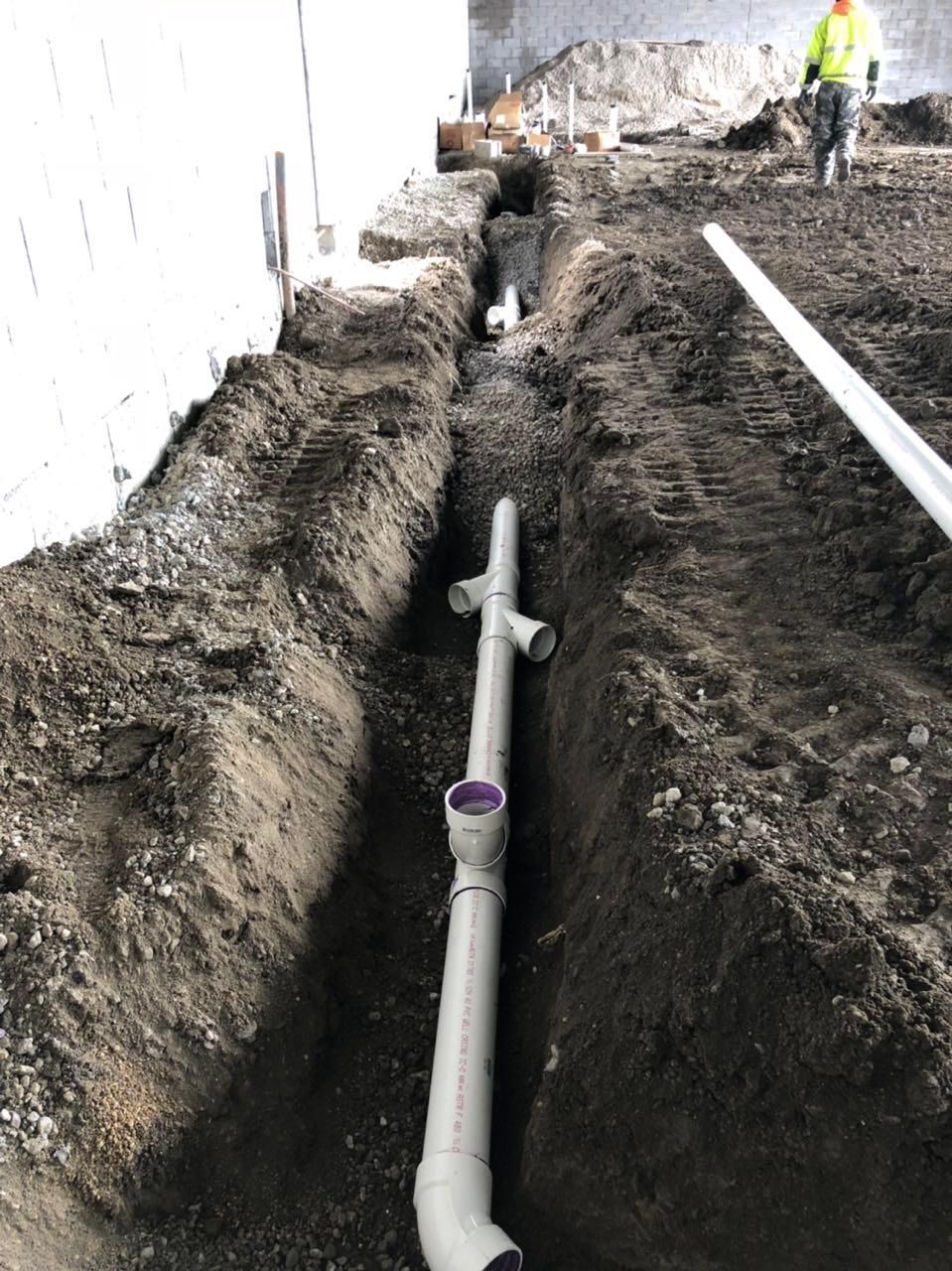 Trench with installed PVC piping near a building foundation. A worker walks in the background.