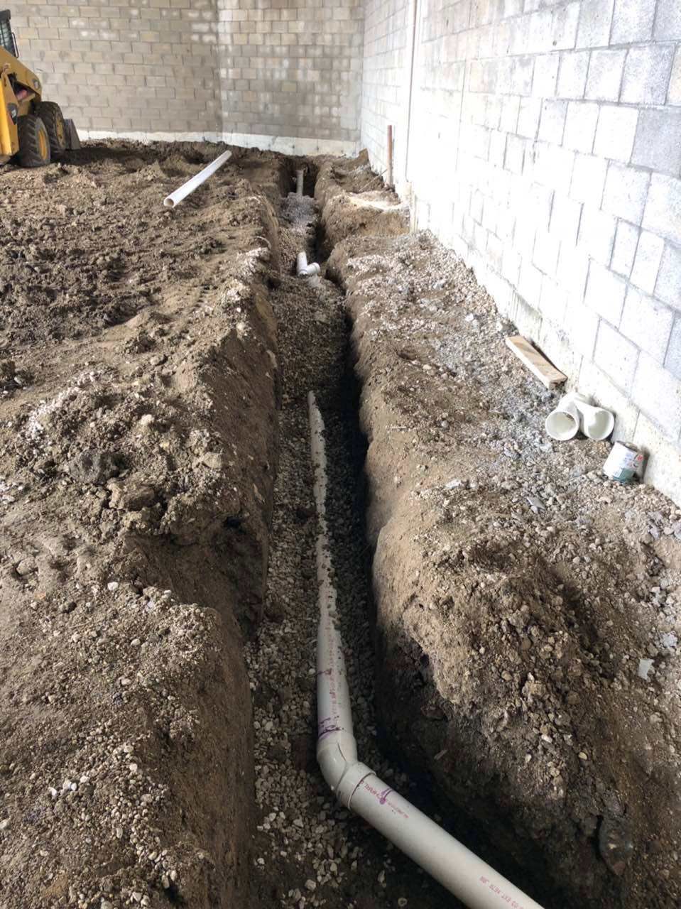 Trenches dug in dirt floor with PVC pipes, inside a building under construction.