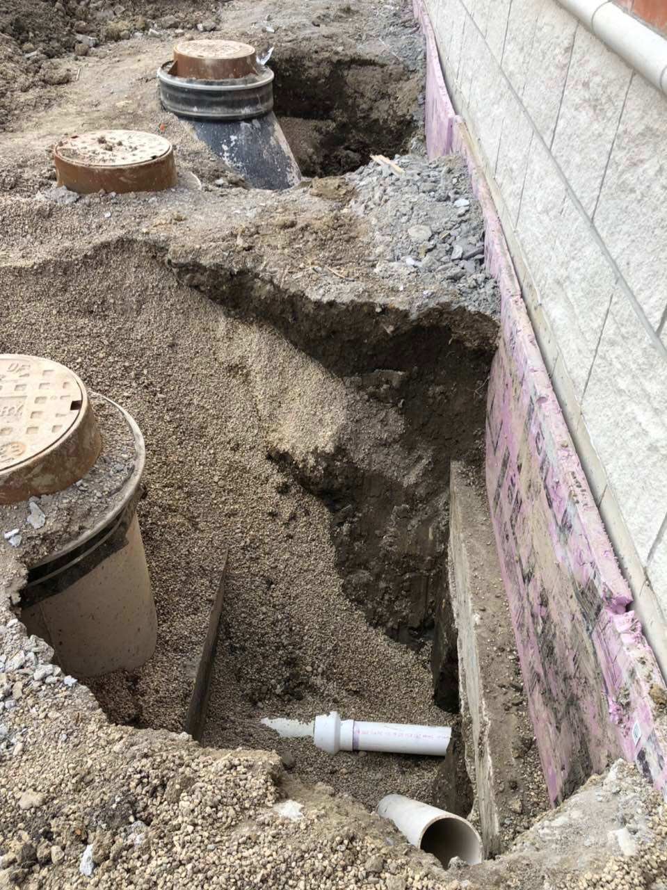 Excavation near a building with gravel, pipes, and access covers. A white pipe is visible.