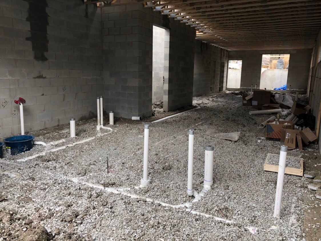 Interior of a building under construction, showing plumbing pipes laid on a gravel floor.
