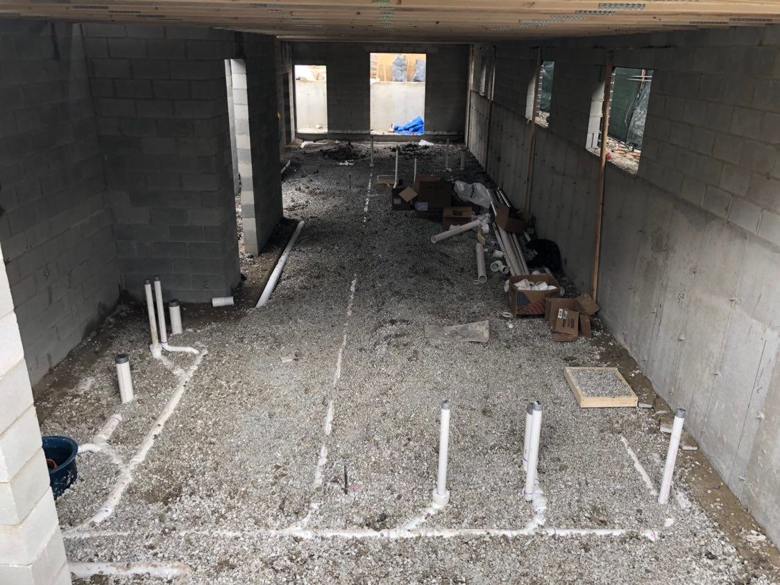 Construction site interior with plumbing pipes laid out on gravel floor. Concrete walls and a doorway visible.