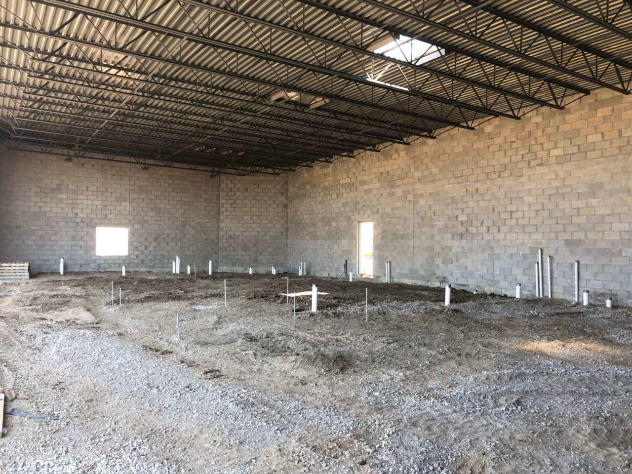 Inside view of a large, unfinished building with concrete block walls, metal roof structure, and exposed plumbing.