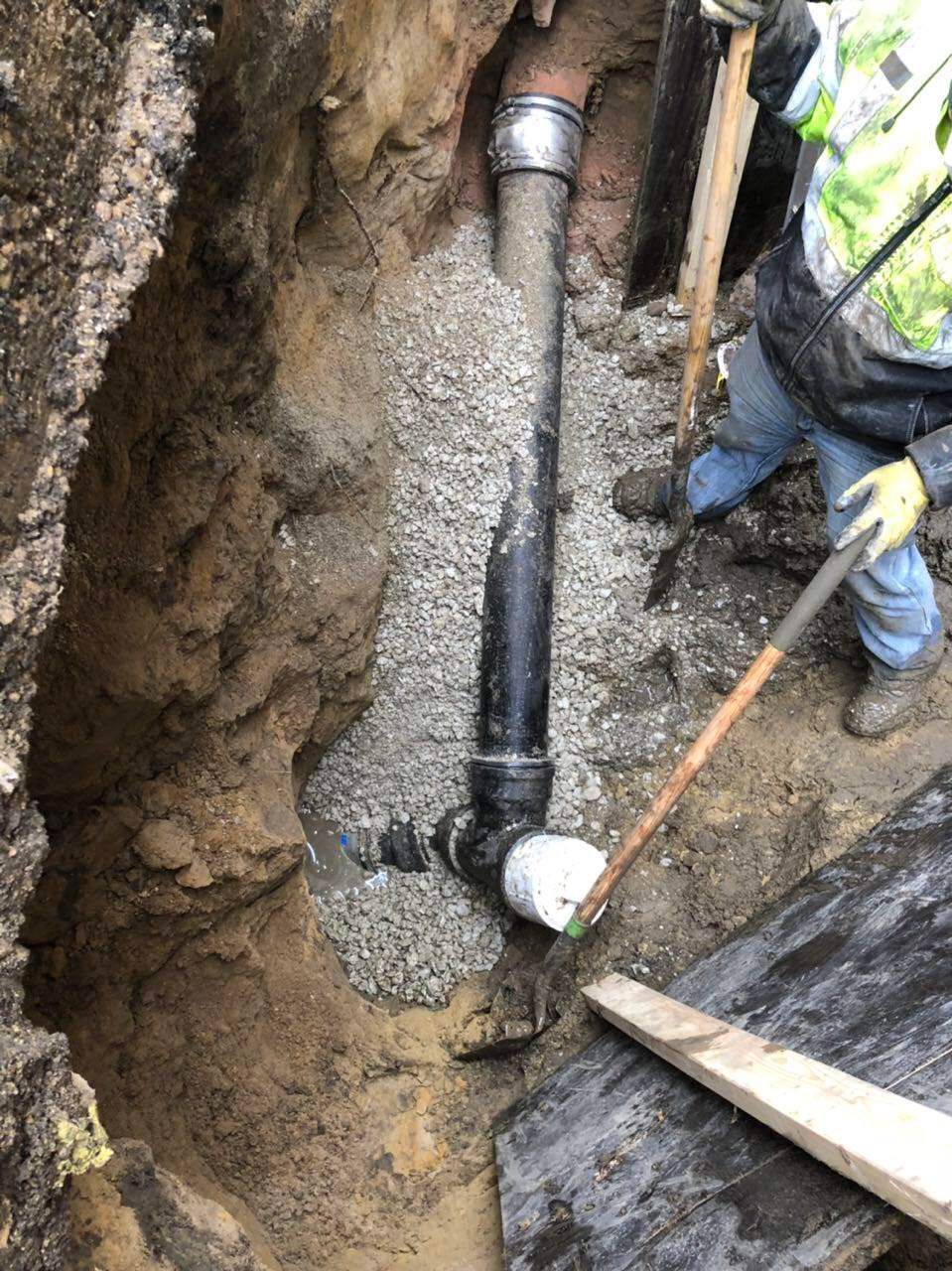 Plumber in trench, replacing pipe. Black pipe, gravel, brown soil, person in high-vis vest.