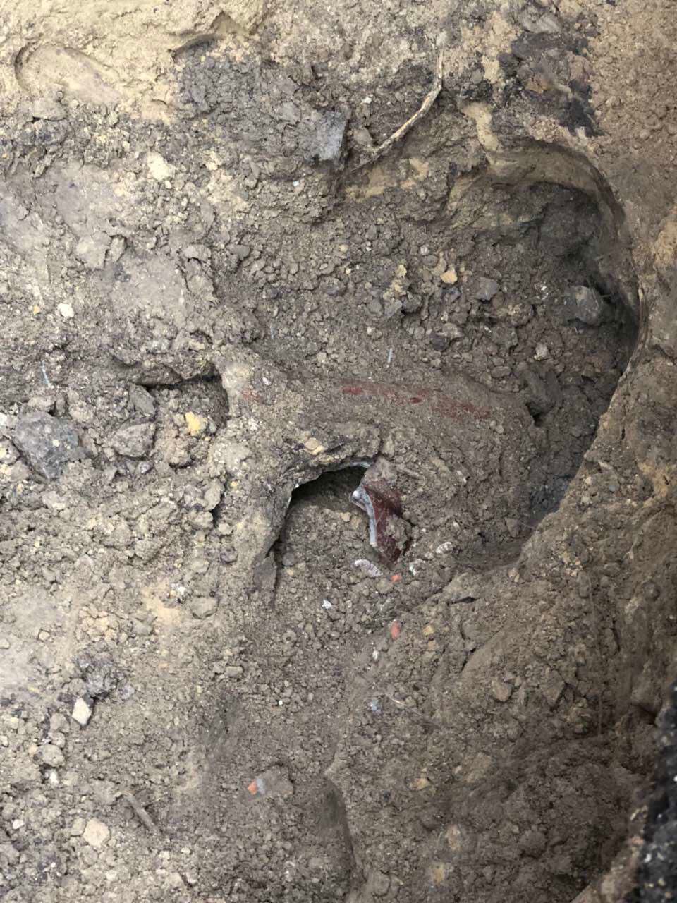 Hole in the ground with a corroded brown pipe visible, surrounded by dirt and debris.