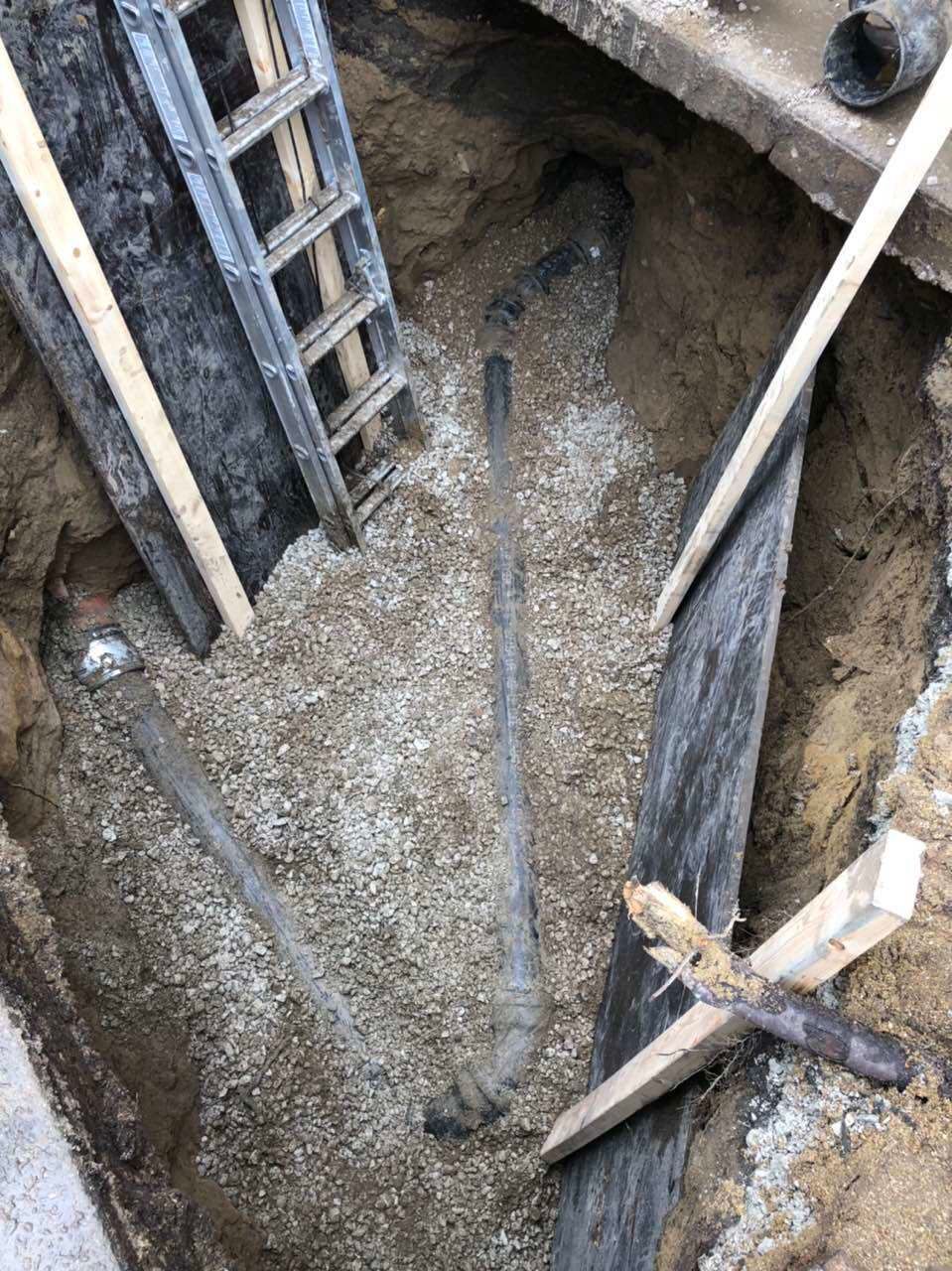 Hole in ground with gravel, tools, ladder and wooden planks.