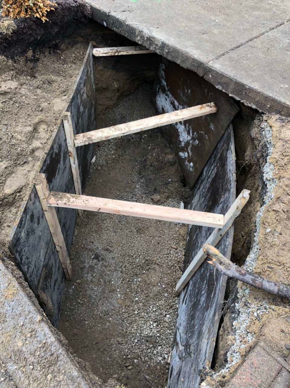 Open trench in concrete sidewalk, braced with wooden supports.