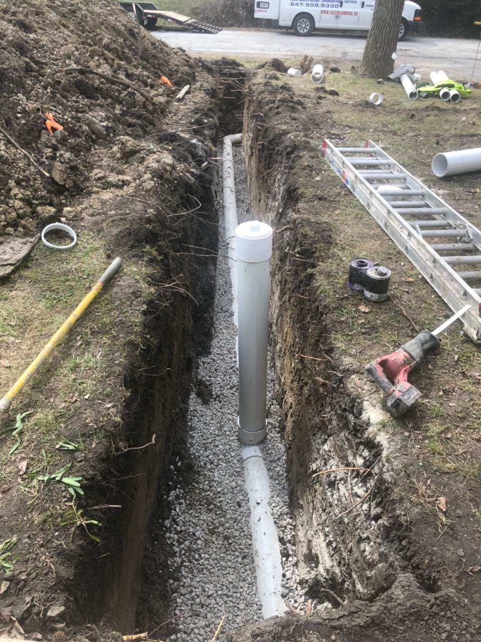 Trench with gray PVC pipe surrounded by gravel. Tools and a ladder are nearby.