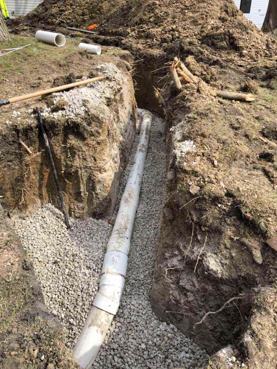 A trench dug in the ground with white piping and gravel, likely for drainage.