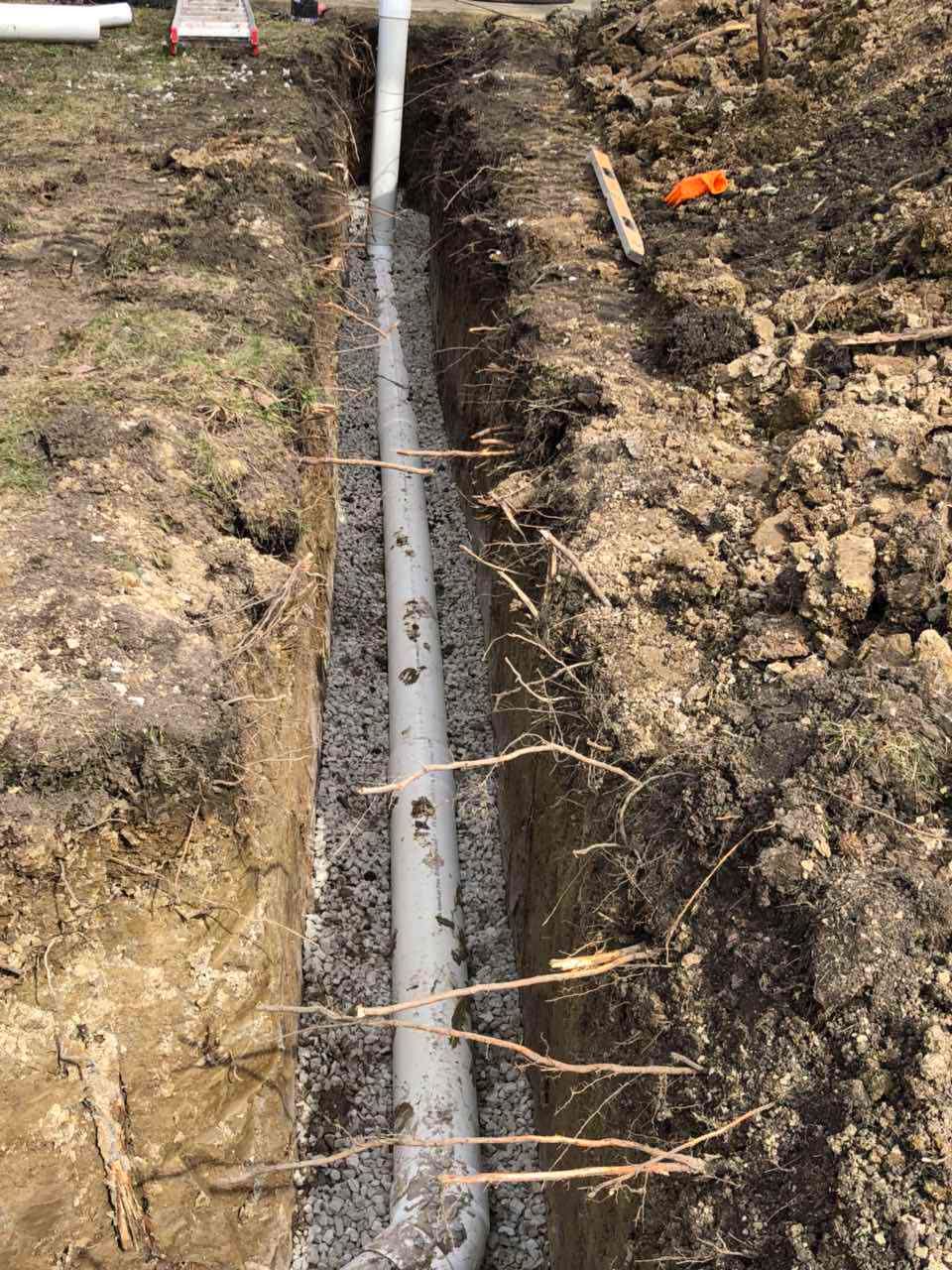 A trench holds a white pipe surrounded by gravel. The ground is dirt and grass, a construction setting.