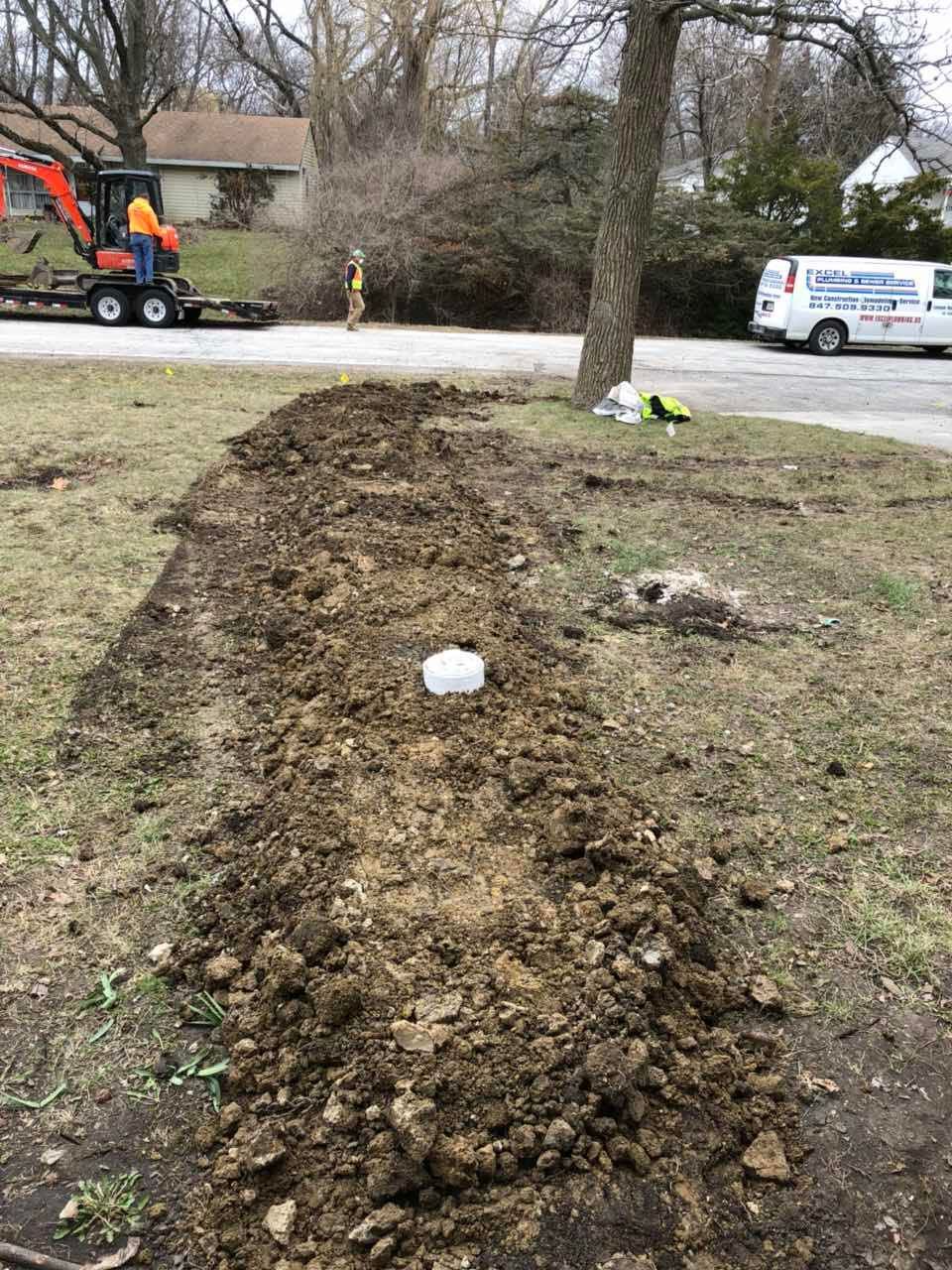 Trench dug in yard with white pipe visible; mini excavator and van in the background, possibly utility work.
