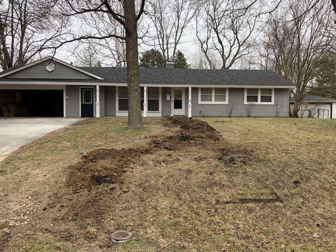 Grey ranch-style house with garage, brown earth patch in front yard, leafless trees.