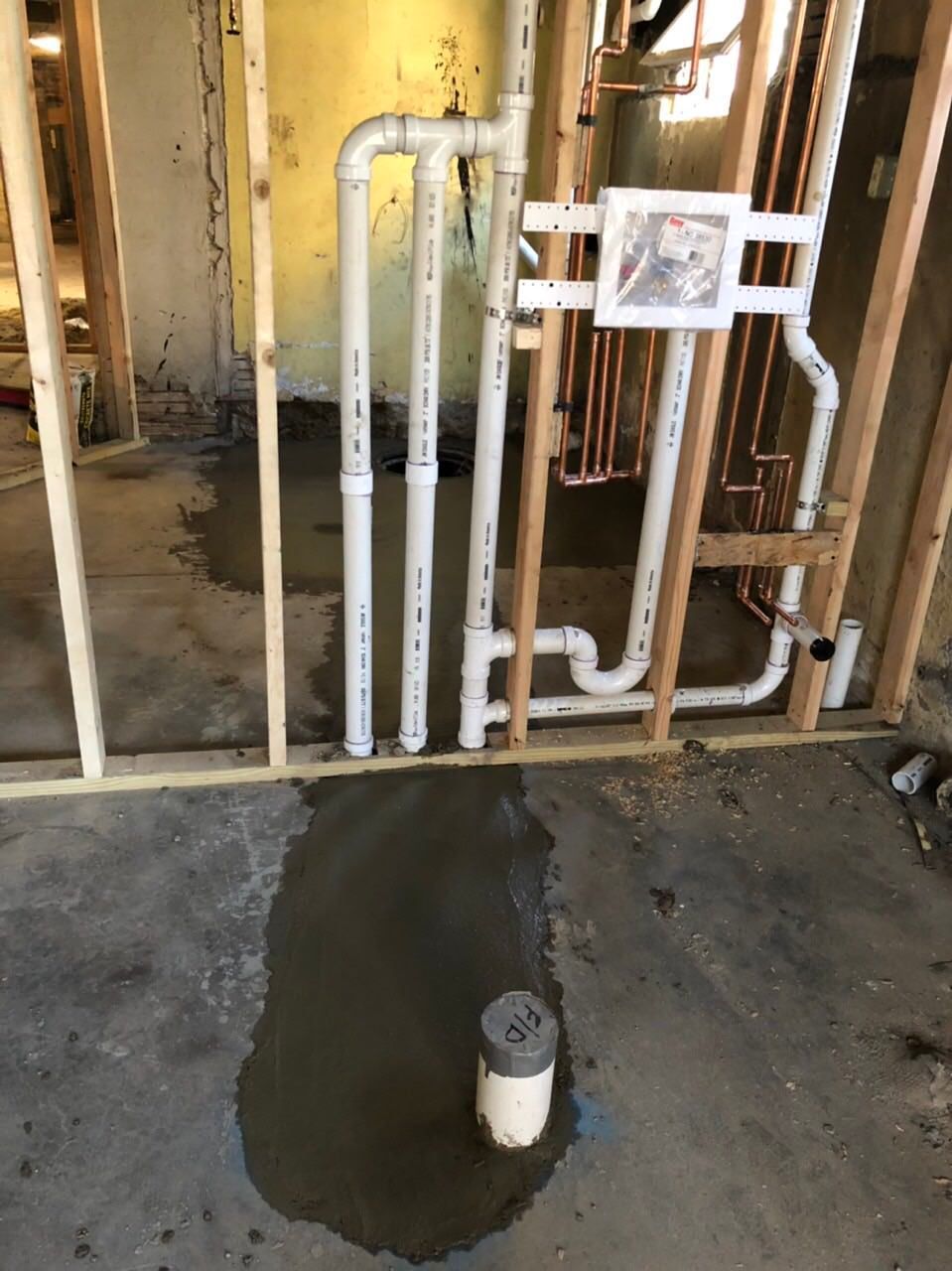 Plumbing pipes and a drain installed in a partially finished wall. Standing water on the concrete floor.