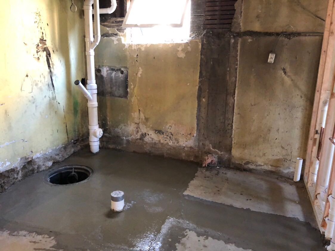 A basement with a freshly poured concrete floor.  A black drain, plumbing, and a small window are visible.