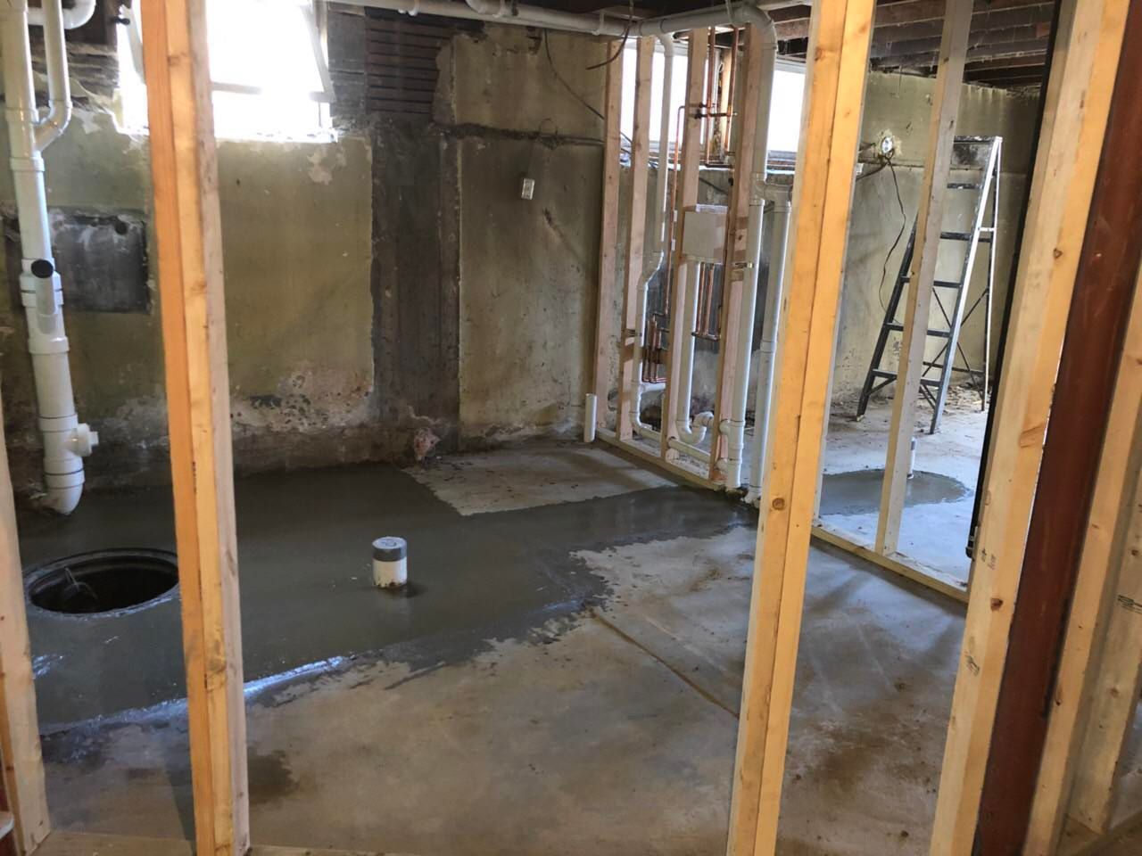 Basement under construction; new concrete floor, wood framing, plumbing pipes visible.