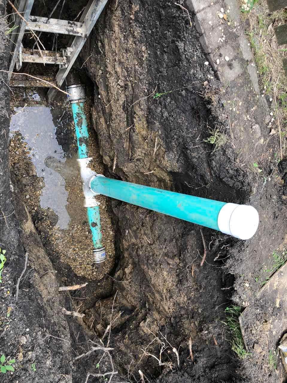 Trench with a green PVC pipe and T-fitting. The trench contains water. Dirt surrounds the pipe.