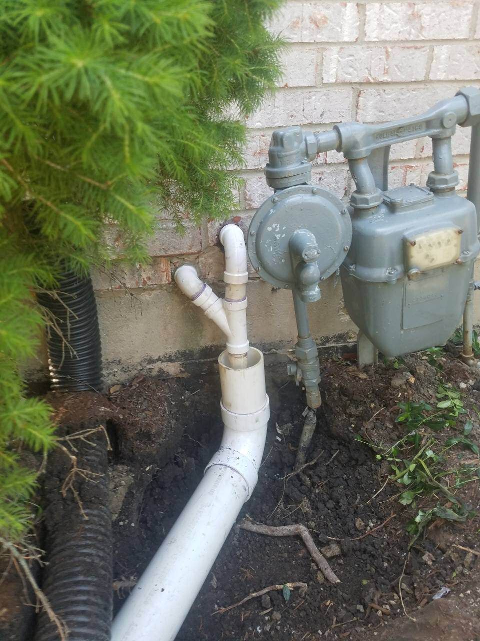 White PVC pipes connected to a gas meter on a brick building with a pine tree to the left and exposed dirt.