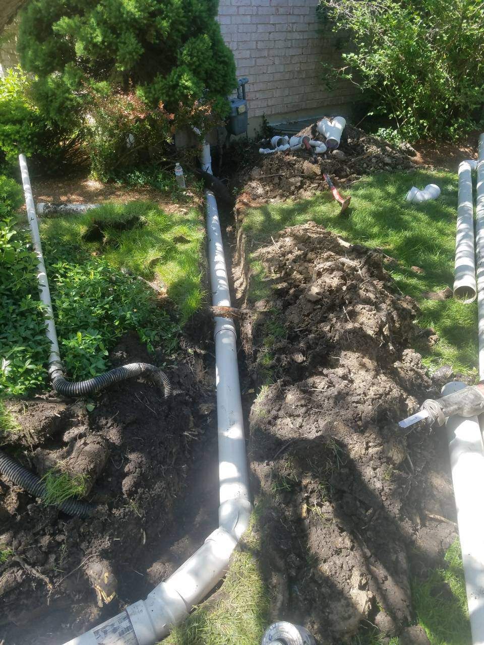 Trench dug in yard with grey pipes laid inside, near green bushes and a brick building.