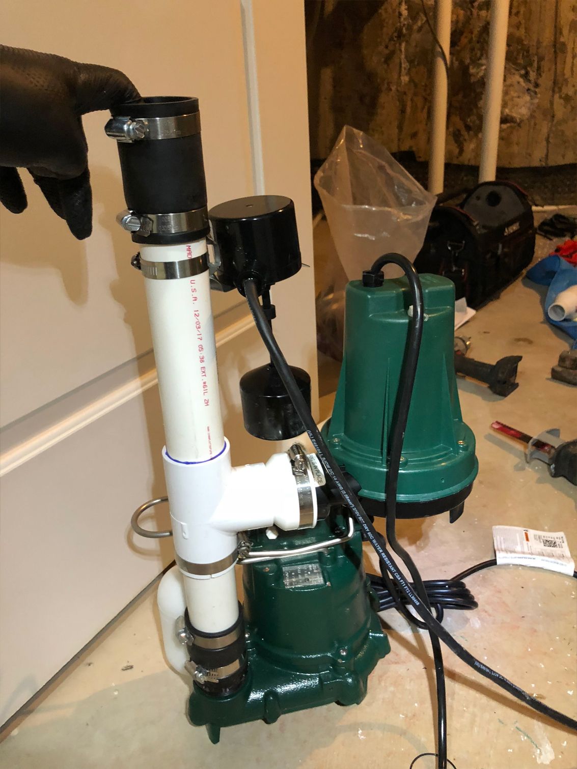 Green sump pump with black float switch and vertical white PVC pipe in a basement.