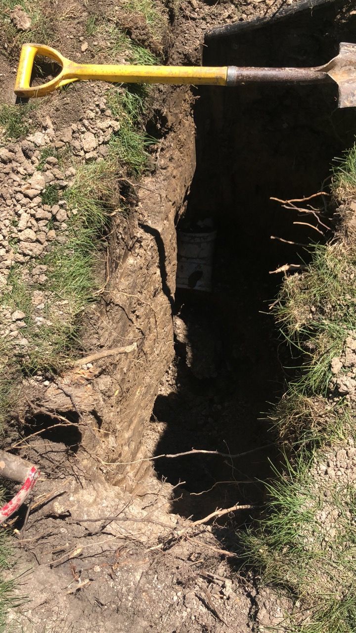 Trench dug into the ground with a yellow shovel resting at the top. Roots and soil visible.