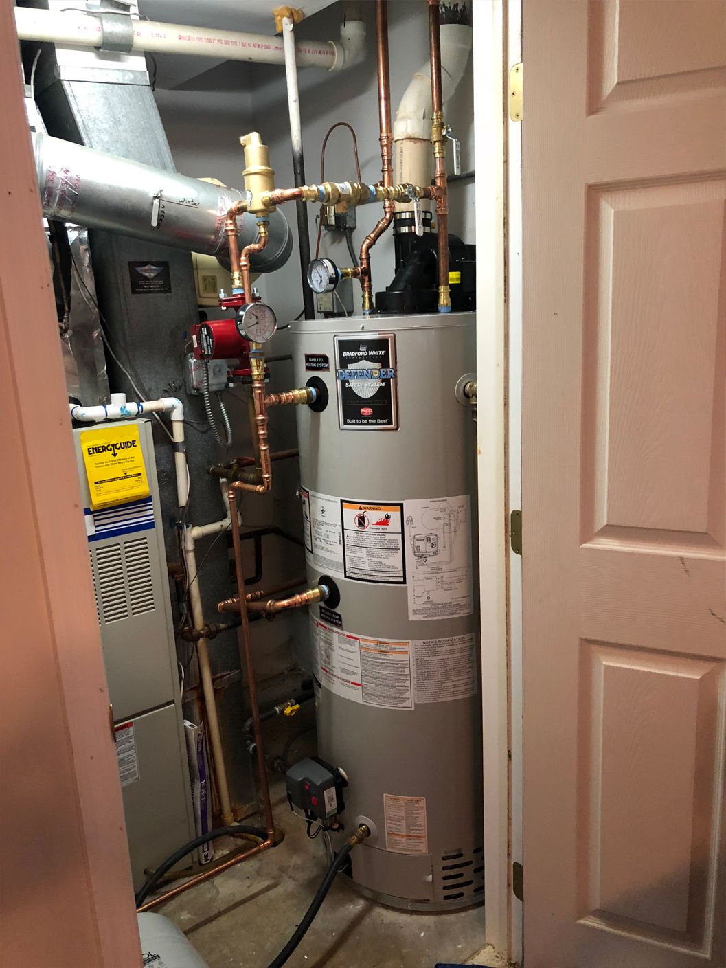 Water heater and associated copper pipes in a utility closet with a furnace, door, and a red circulating pump.