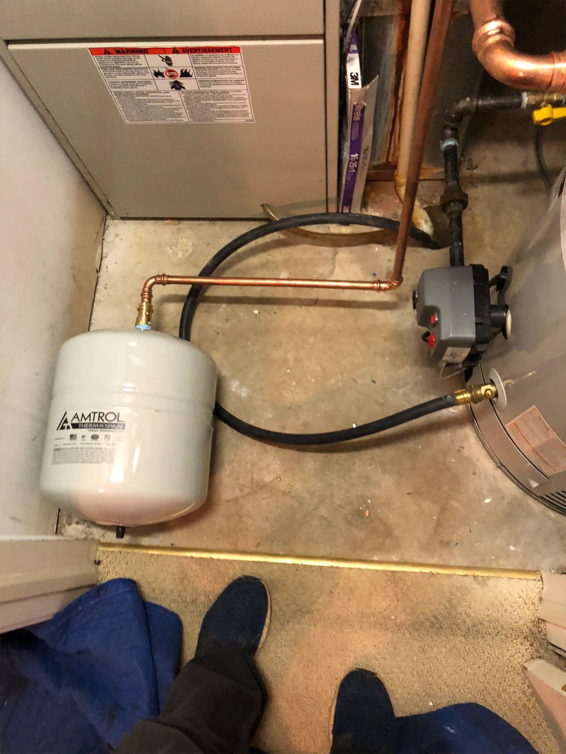 White expansion tank and heating pump plumbed with copper and black tubing near a furnace.