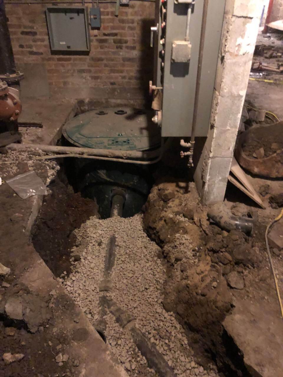 Basement with open pit around a dark cylindrical object and electrical box on a brick wall. Gravel and exposed pipes.