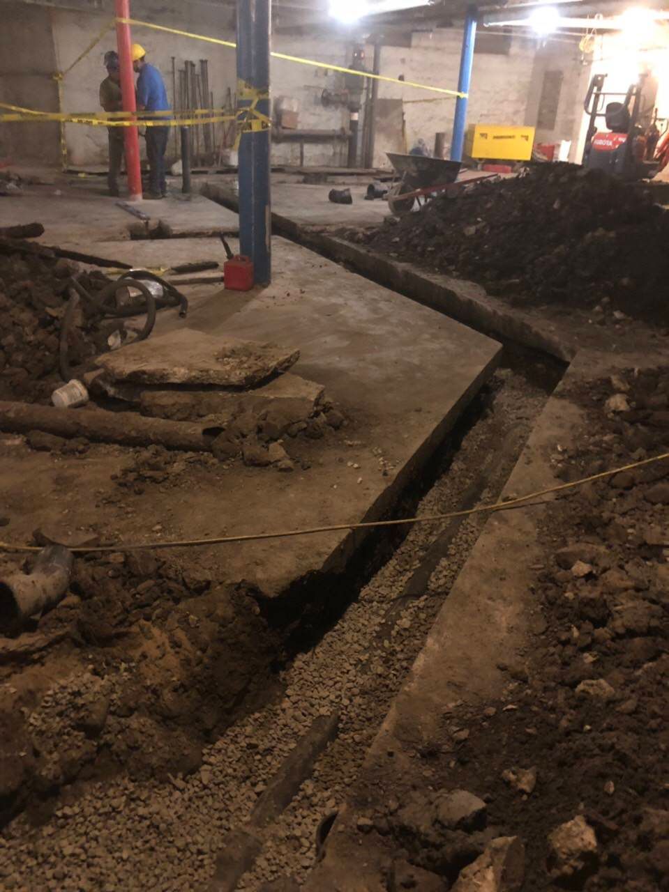 Trench dug in a concrete floor. Workers in hard hats and equipment visible in a dimly lit basement.