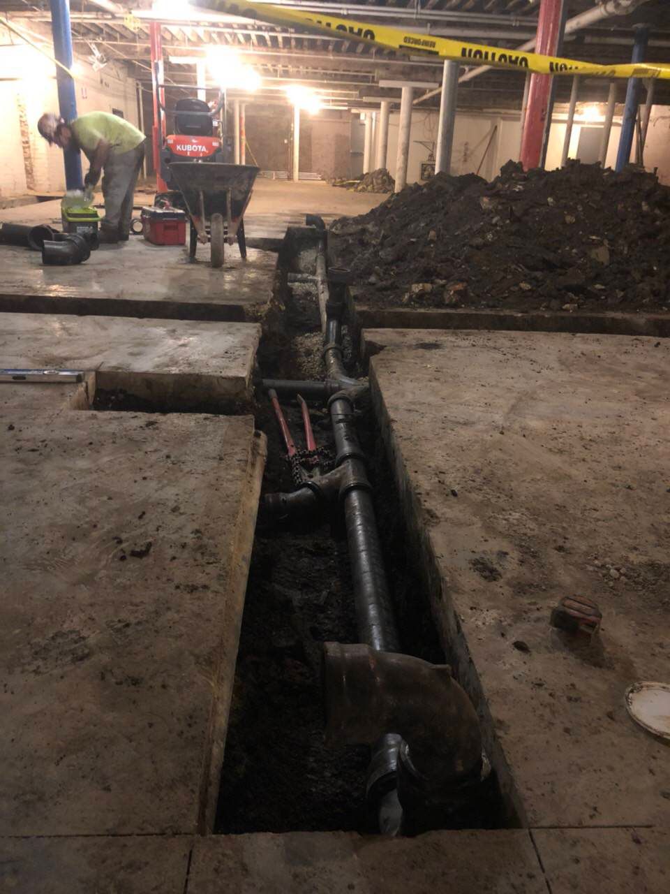 A worker repairs underground pipes in a concrete basement. Dirt, tools, and caution tape are visible.