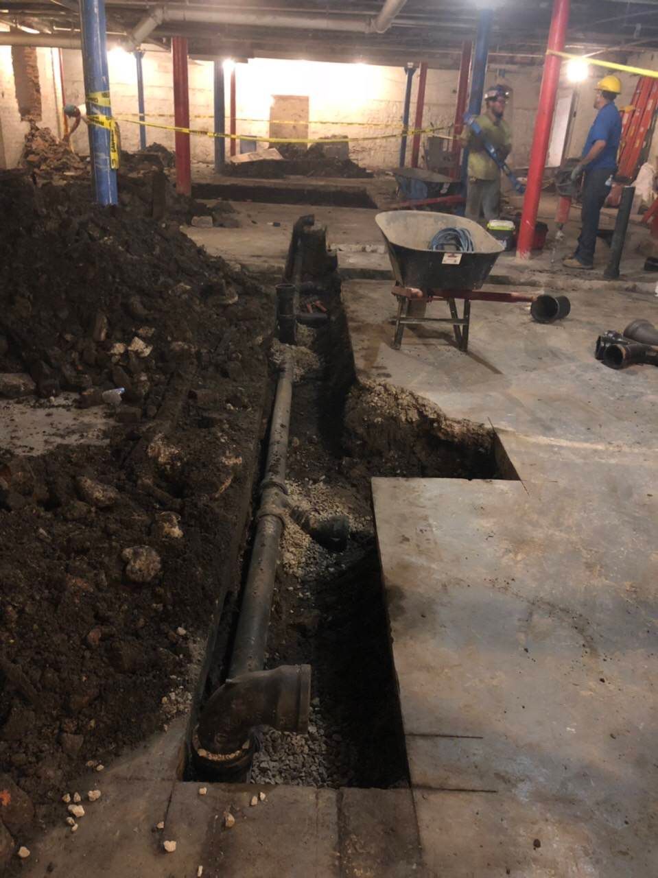 Basement excavation: a trench next to a pipe. Workers, dirt pile, concrete floor.
