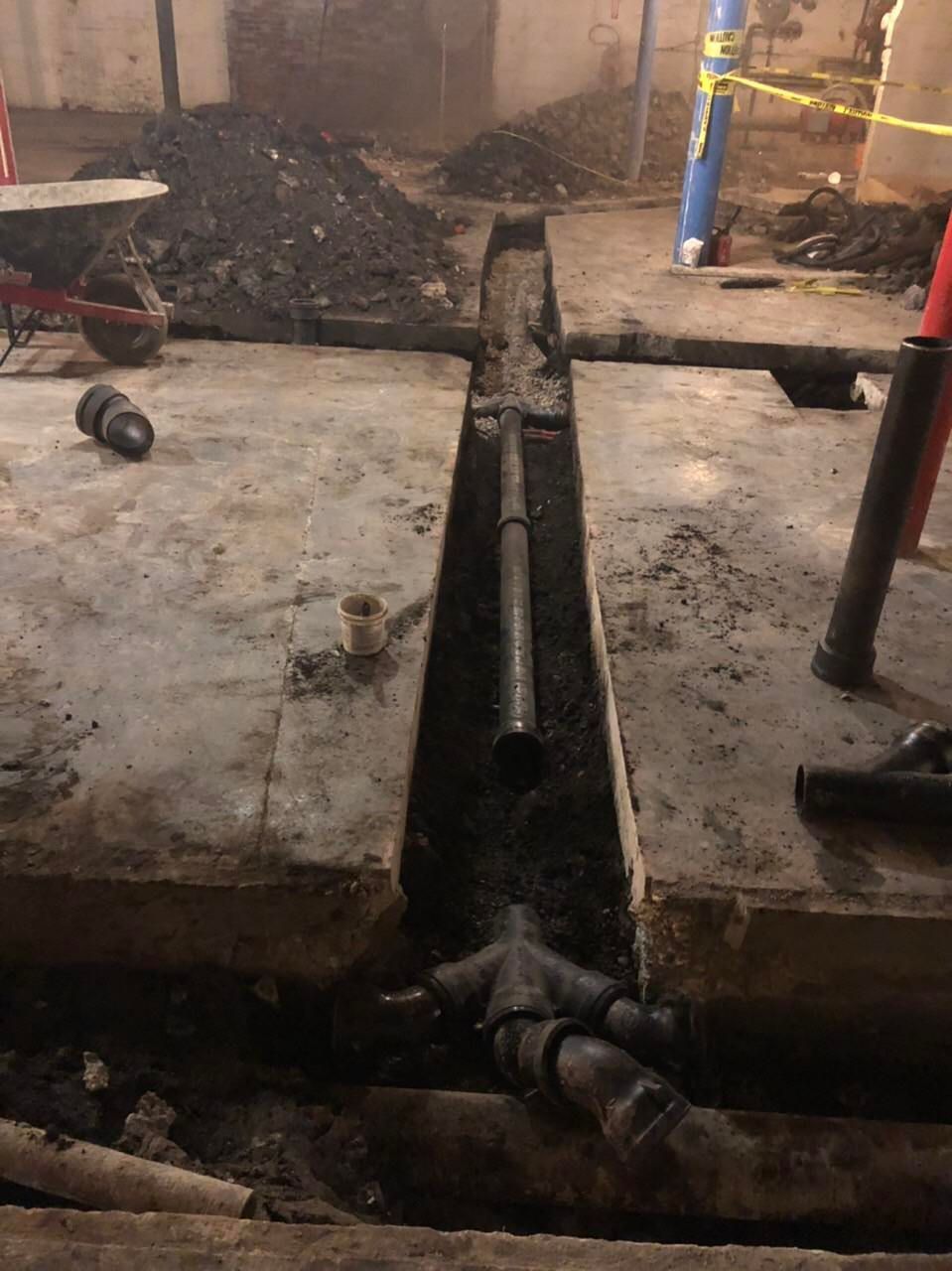 Trench cut in concrete floor, revealing plumbing pipes. Dirt pile and tools in a construction setting.