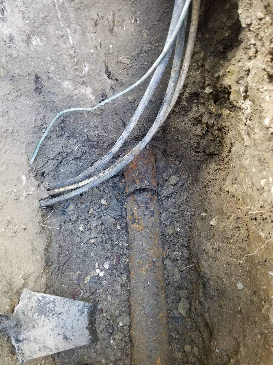 Trench in dirt reveals rusted metal pipe and electrical wires; a shovel rests nearby.