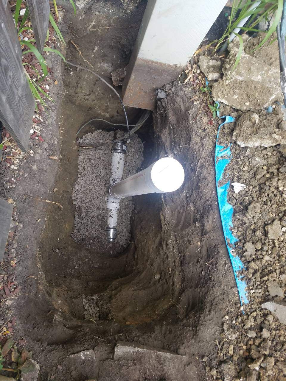 Trench dug in the ground with a vertical white pipe and connected components near a wooden structure.