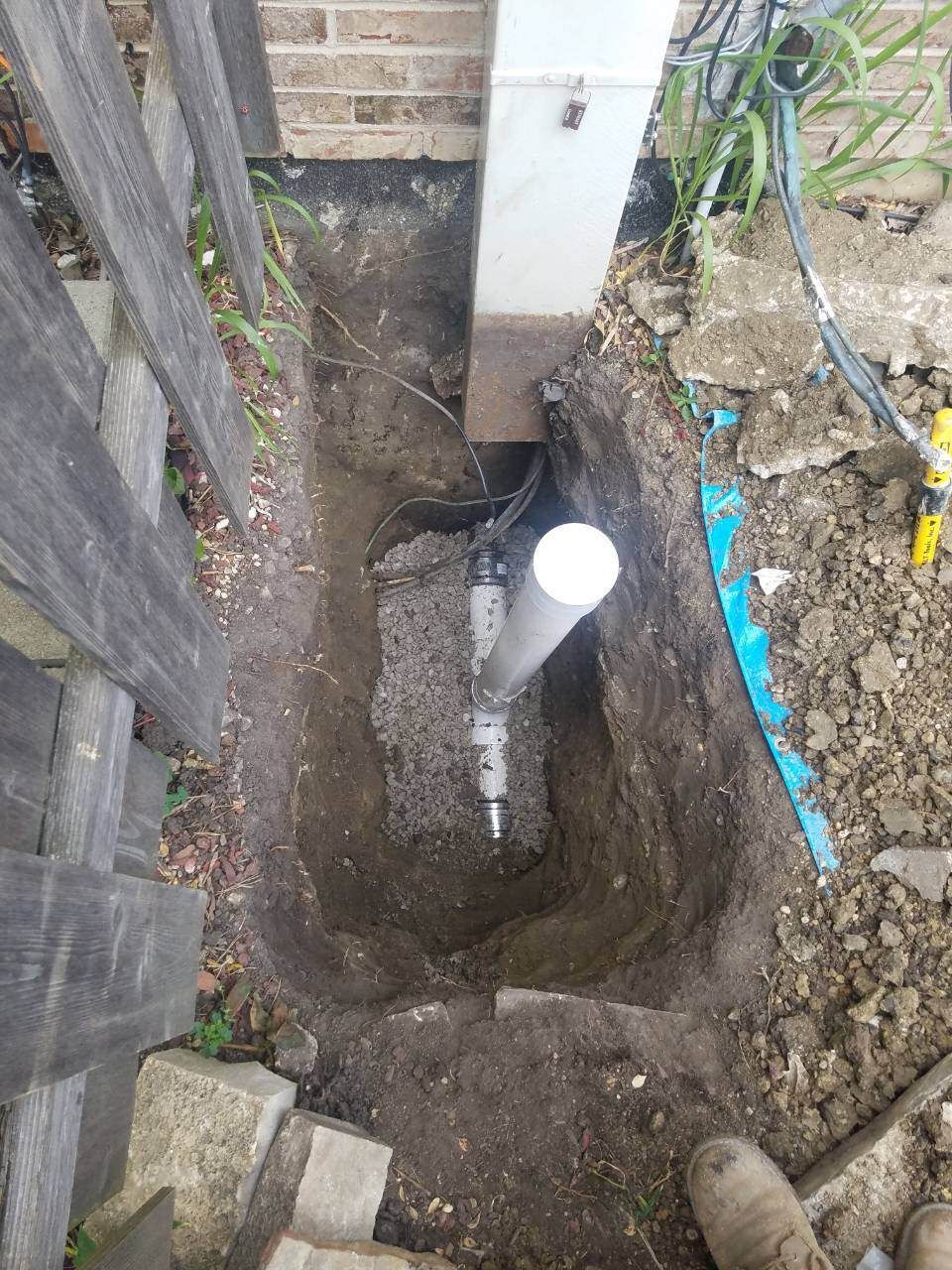 Dug trench next to a fence and a wooden post holding a PVC pipe and related plumbing.