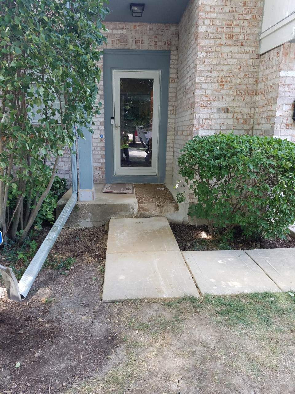 Exterior view of a home's entrance: clear glass door, concrete path, brick facade, bushes, and a downspout.