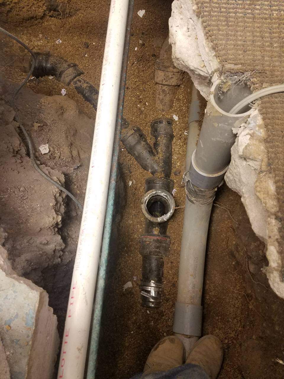Plumbing pipes exposed in a trench. Black, gray, and white pipes with a coupling. Dirt and concrete present.