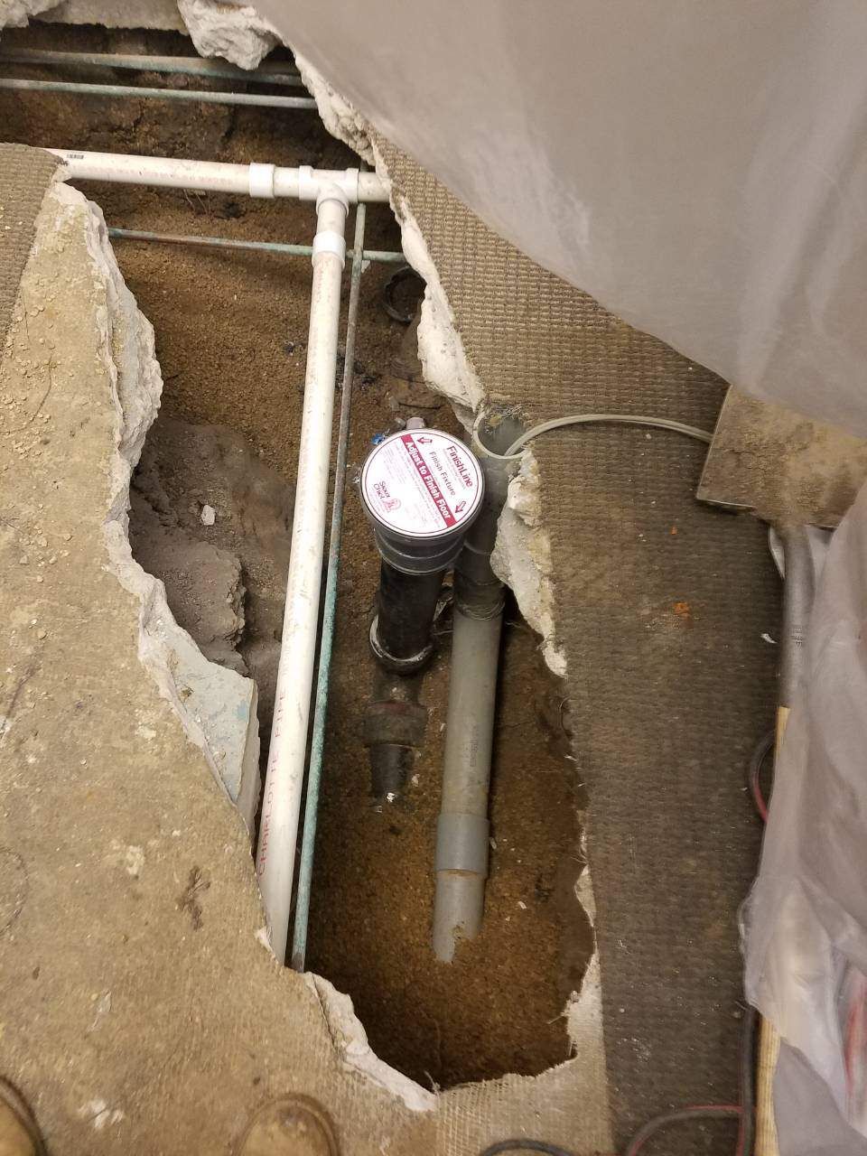 Hole in floor reveals plumbing pipes. White, gray and black pipes, in a concrete and carpet setting.